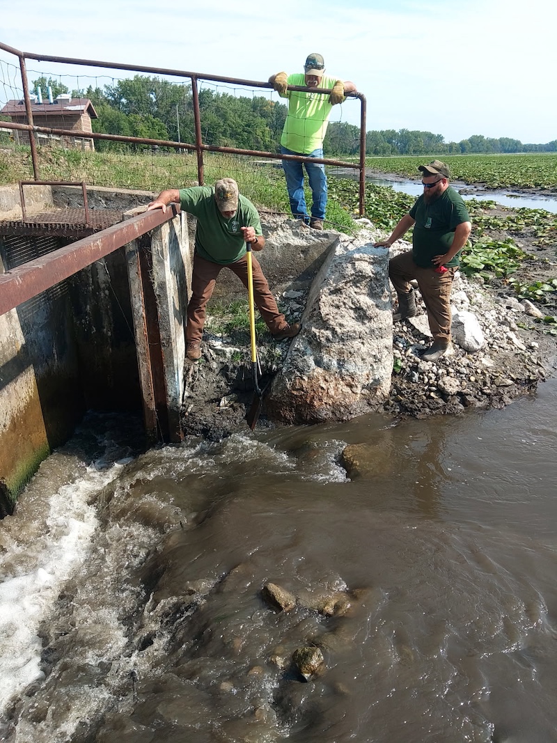 Three men stand next to a deep ditch that allows extra water to run-off from a lake. One of the men holds a shovel near the water flowing through the channel. In the background is the shoreline and lily pads filling. Trees are against a bright partly cloudy sky.