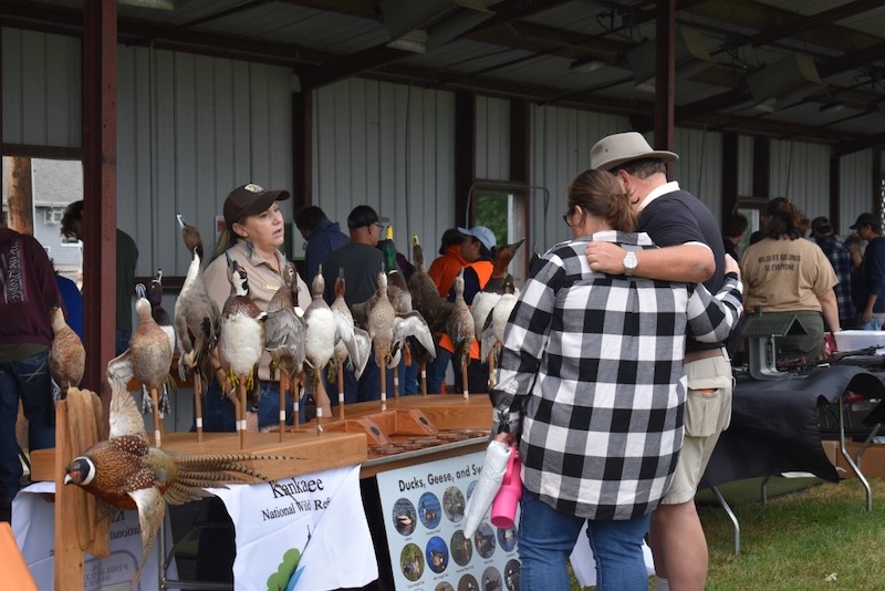 A woman and man stand together in front of a long table set up will identifying information for different species of wild waterfowl. In the background is a metal pavilion with a partial roof. People are standing in the pavilion looking at different displays and conversing.