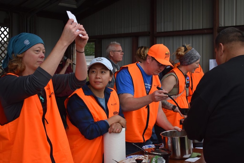 A row of people wearing blaze orange gear ladle out food on to paper bowls from stainless steel pots. One individual holds up some card with both hands above his head.