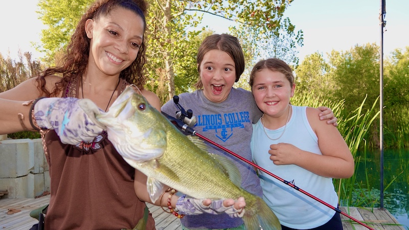 On a wooden dock over a pond a woman holds up a large green and white fish while two excited young girls stand next to the woman. One of the girls holds the fishing rod and reel. 