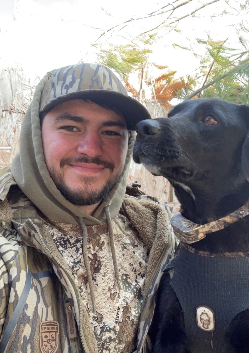 A portrait of a man wearing camouflage gear and his black Labrador retriever. The dog has a camouflage collar. In the background is tan and green vegetation.