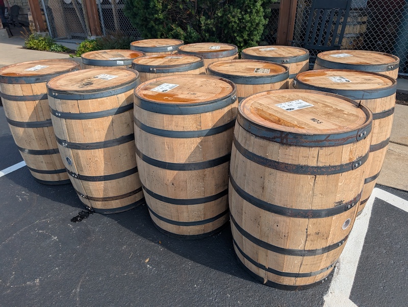 A group of twelve barrels standing upright on the edge of a black asphalt parking lot.
