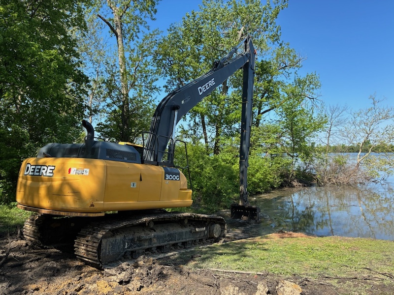 A large excavator pulls a bucket of earth towards the shoreline of a lake. Trees fill the shoreline in the background. A bright blue sky is overhead.