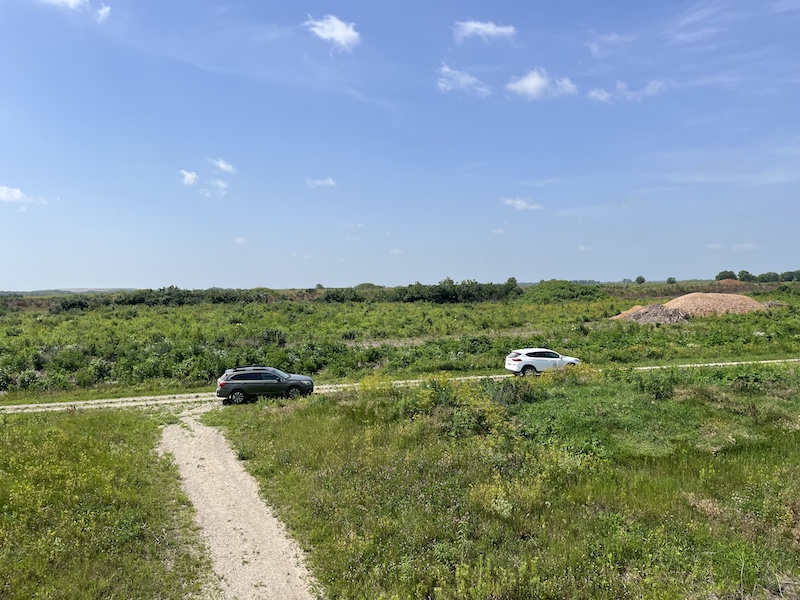 A bright blue sky is above a view overlooking a prairie.  Two SUV cars are parked along a gravel road. A path cuts through the prairie towards the foreground.
