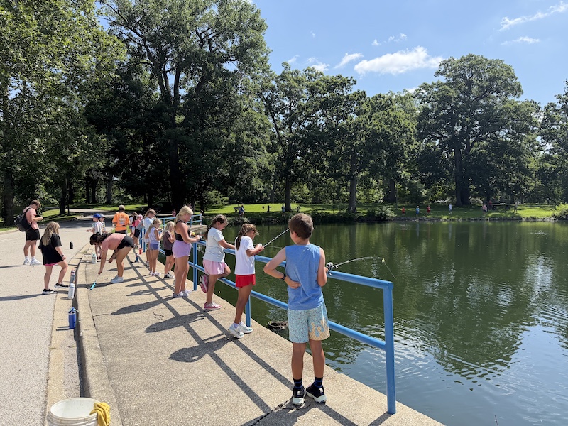 A group of children stand on a concrete bridge overlooking a pond and hold fishing rod and reels. All the children are attempting to catch fish. In the background sunlight is dappled on mowed park lawn in between the shade of tall beautiful trees. A bright partly cloudy sky is overhead.