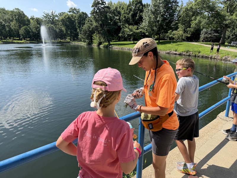 A woman takes a small green fish off the hook while a young girl holds the fishing rod and reel the fish is attached to. In the background are more children and adults fishing off a concrete bridge over a pond.