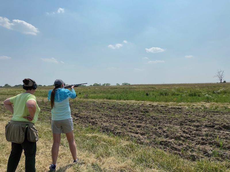 Two women stand near a grassland. One woman aims her shotgun towards the field. The woman behind her stands with her hands on her hips and guides her through the process. A bright blue sky is overhead.