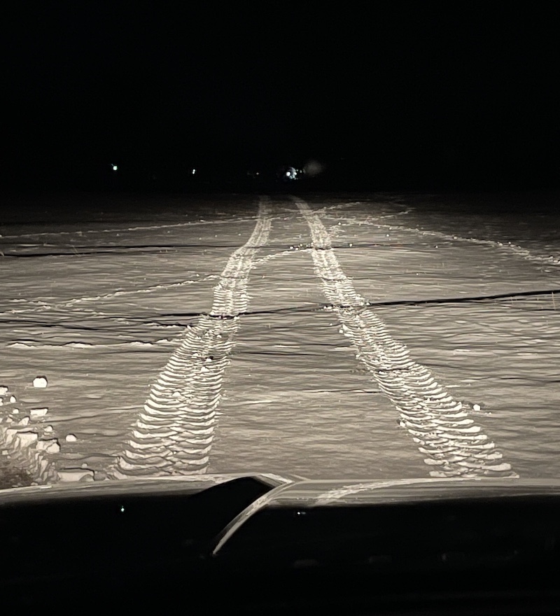 A nighttime view of a snowy field. Headlights from a pickup truck illuminate a field and reveal tracks of a vehicle moving out into the field. Lights can be seen in the direction of the tracks in the distance in the middle of the field.