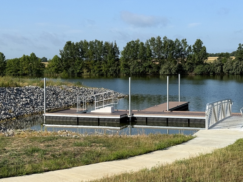A dock on a freshwater lake. In the background is a rocky shoreline. A trees fill the shoreline in the distance.