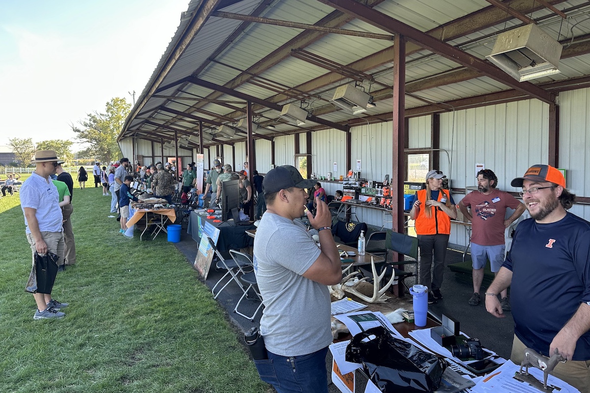 During a summer day under a bright blue sky a row of tables are lined up in front of a partially roofed long metal pavilion. People stand on either side of the tables and engage in conversation. Various items and hunting gear rest on the tables and on the shelves behind the tables.