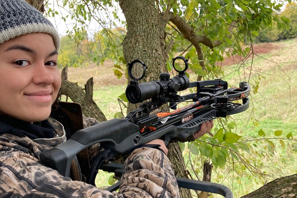 A hunter holds a crossbow up in preparation for aiming while sitting in a tree-stand. In the background is a grassy field. Trees with autumn foliage border the field in the distance.