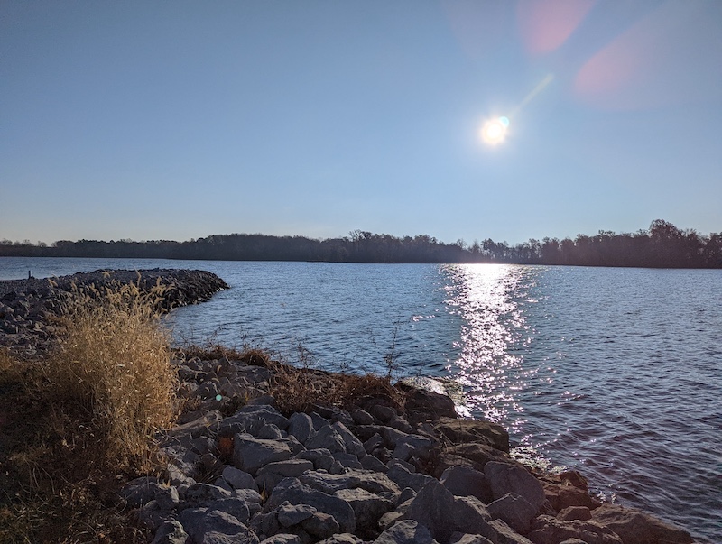 Bright blue sunny sky is reflected upon the surface of a wavy lake. In the foreground is a rocky lake shore. Trees form a horizon line in the background silhouetted by the bright sun.