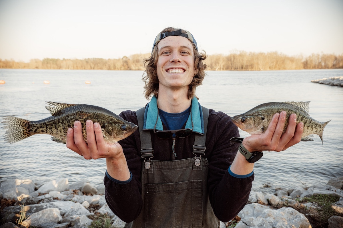With one black and tan speckled fish in each hand, a researcher stands on the bank of a fresh water lake with a big smile. In the background trees fill the lake shore and form a horizon line against a bright blue sky.