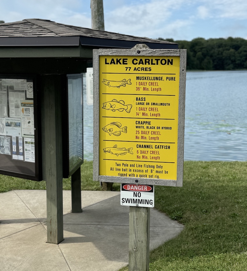 A yellow sign secured to a wooden post is installed on the grassy bank of a freshwater lake. The sign has line illustrations of four different fish including muskellunge, bass, crappie and channel catfish. Each fish has the legal limit and length that an angler may take each day. In the background is a kiosk with posters and more information.