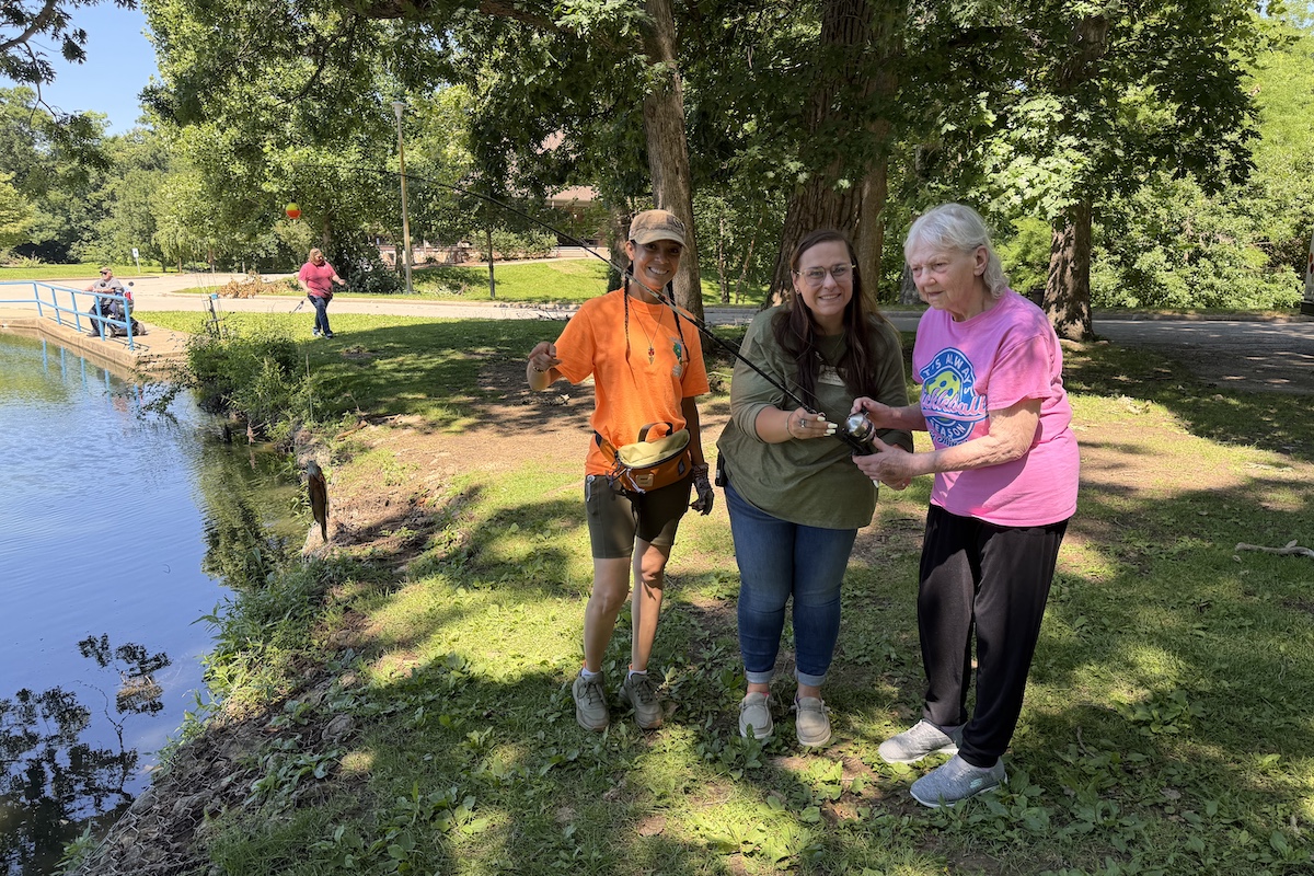 An elderly woman holds a fishing rod and reel with a fish caught on a hook attached to the fishing line. To women stand next to her. One of the adjacent women helps the elderly woman hold up the fishing rod and reel. In the background is a pond in a park with trees and a quiet street nearby. Two people fish from the pond bank.