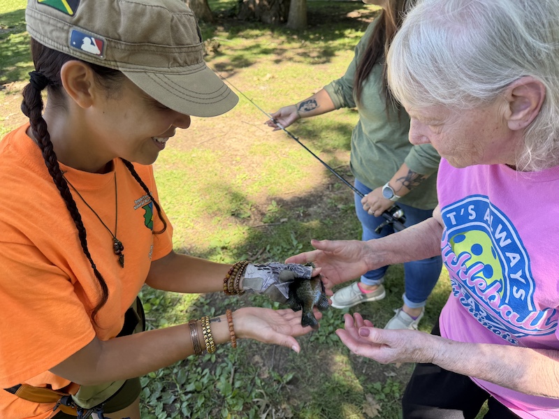 A woman places a fish in the hands of an elderly woman. In the background is another woman holding the fishing rod and reel. 