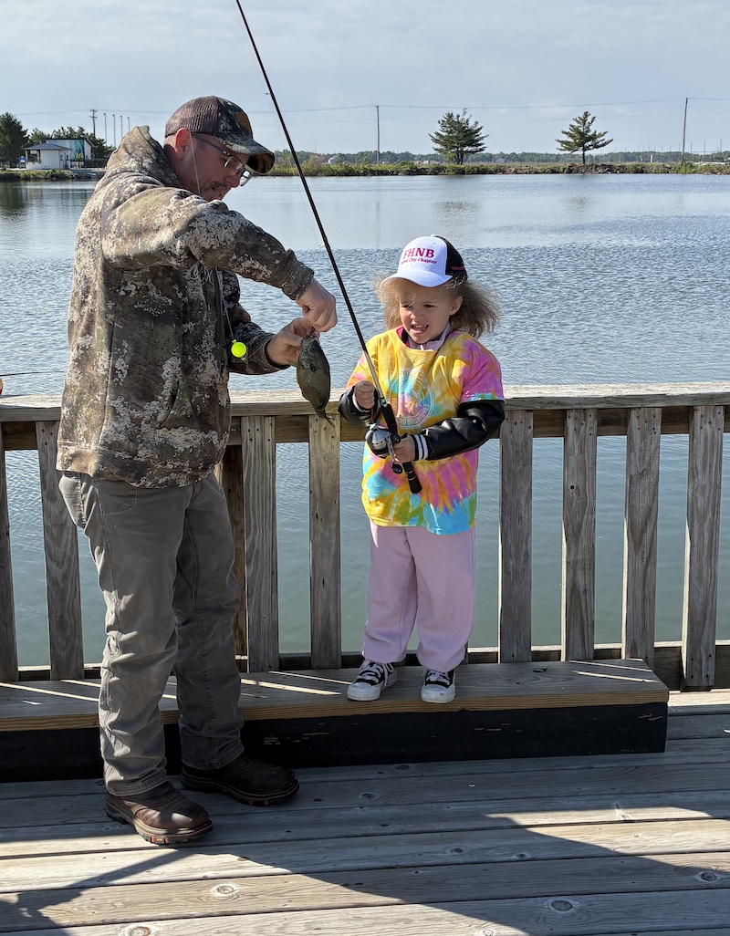 A man works to take a small green fish off a hook successfully caught by a young girl. The man and girl are standing on a wooden pier overlooking a pond.
