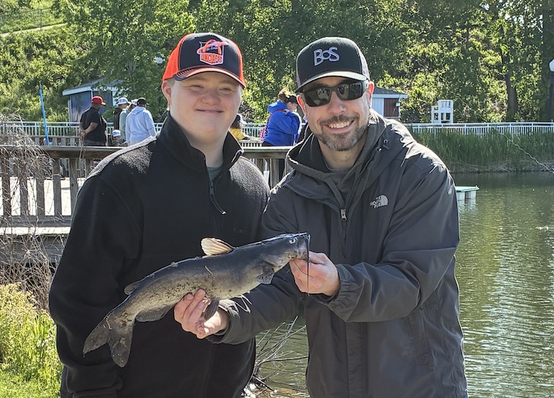A man stands next to a teenager and holds up a medium sized black and tan catfish. IN the background is wooden pier overlooking a pond. Many people are fishing from the pond.
