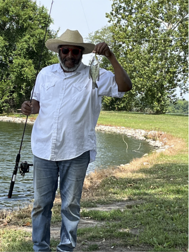 An African American man wearing a hat, sunglasses and short-sleeved, button down shirt poses with a fish that he caught.