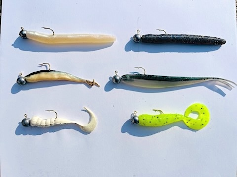 An arrangement of six fishing hooks with plastic "worms" rest on white paper.
