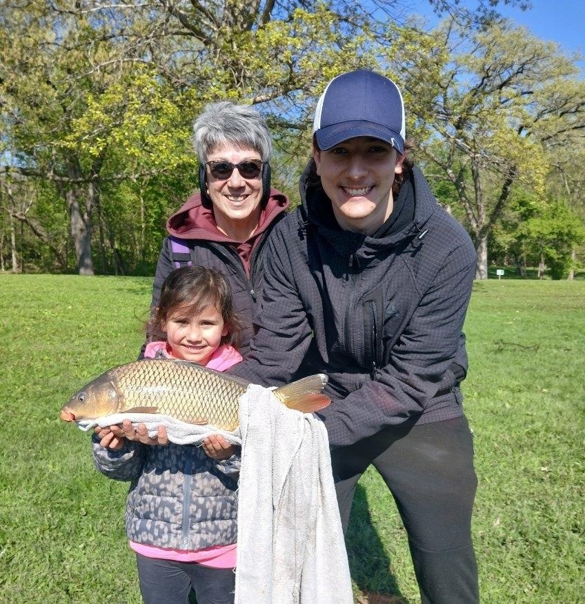 A young girl holds up a large tan fish while a young man next to her helps her hold up the fish. A woman stands behind the young girl. In the background is a sunny park lawn with trees with spring foliage.