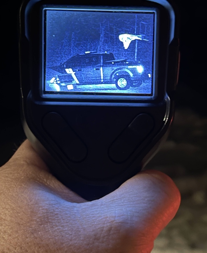 The screen of a night vision device held in the hand of a conservation police officer shows a pickup truck parked next to a sedan. The white shape of an owl flies over the truck.