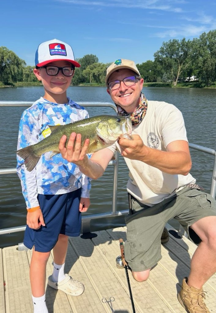 A man holds kneels next to a young boy and holds up a medium size green and silver fish in front of the child. Both individuals are on a dock over a freshwater pond. In the background trees are against a bright blue sky.