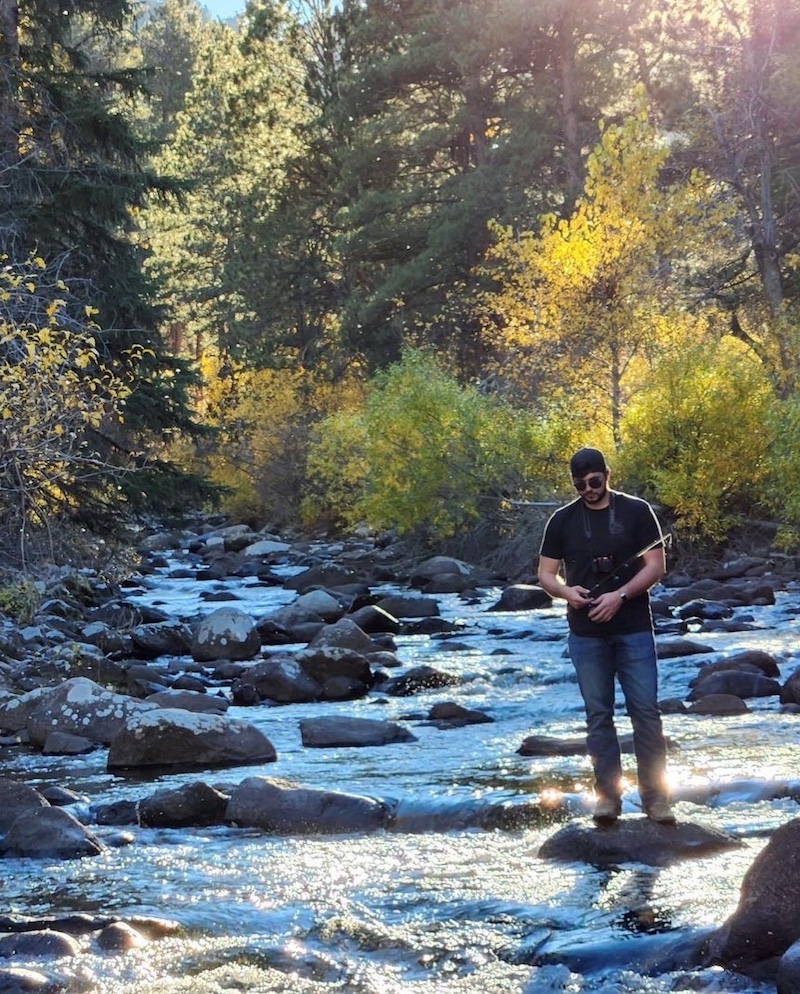 A man stands on a large rock in a river while he holds a fishing rod and reel. In the background is a rocky river. Trees with fall yellow and green foliage are on both sides of the river.