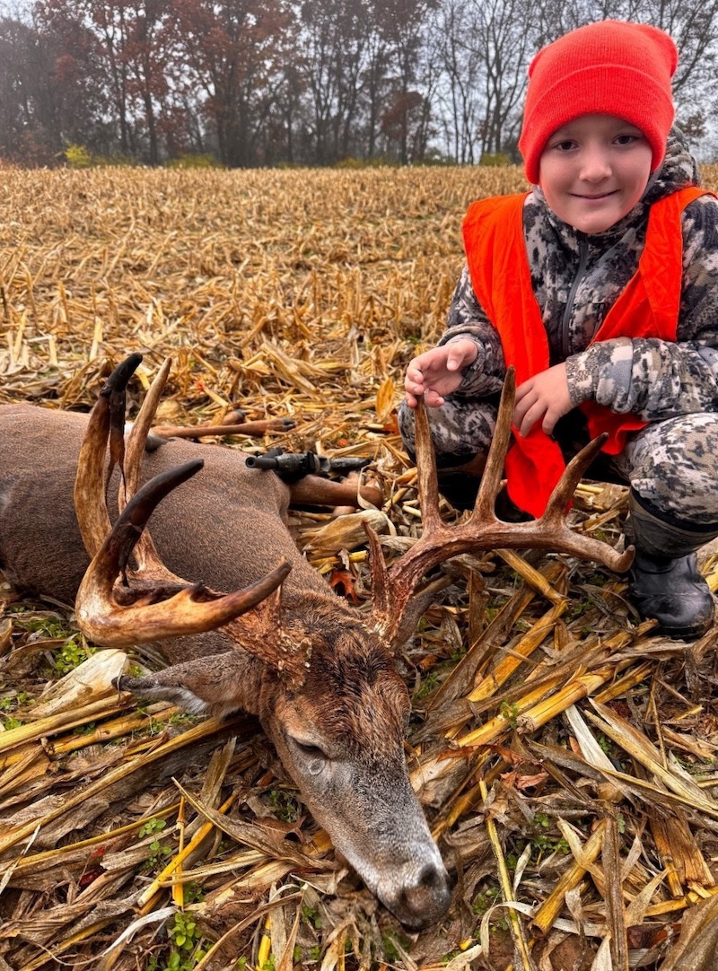 A young adult wears camouflage and blaze orange gear while squatting down in a harvested agricultural field next to a successfully harvested large white-tailed deer. In the background, trees border the field, and an overcast sky can be seen through the tree limbs.
